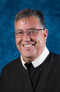 Smiling man in glasses and black clergy robe against a blue backdrop