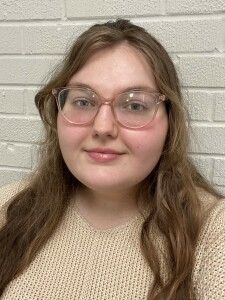 Portrait of a person with glasses and long hair, wearing a beige sweater against a white brick wall