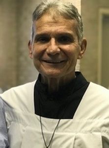 Smiling man in a white apron over a black shirt, standing indoors against a blurred background