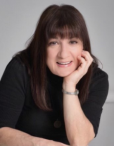 Smiling person with dark hair resting chin on hand, wearing a black top against a gray background.