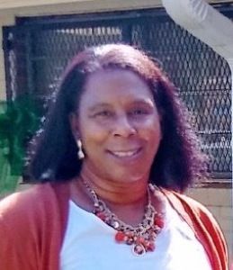 Smiling person outdoors wearing a coral cardigan, white top, and red necklace in front of a metal fence