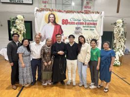 Group of people posing indoors beside a religious banner and statue display at a milestone celebration