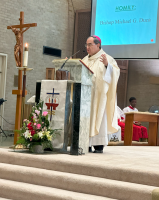 Priest in white vestments speaking at a church lectern beside a crucifix and flowers