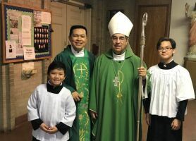 Four people in church vestments, including a bishop in green, posing indoors near a bulletin board.