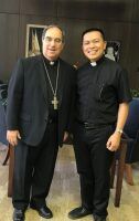 Two men in black clerical clothing standing together indoors, smiling at the camera.