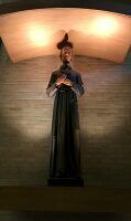 Statue of a robed figure holding an object, lit by warm overhead light against a stone wall.