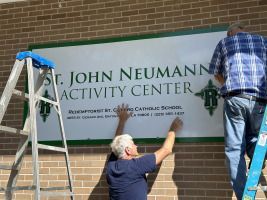 Workers hang a sign reading “St. John Neumann Activity Center” on a brick wall.