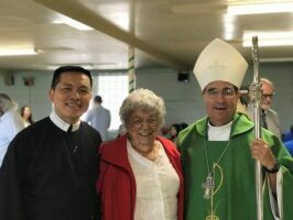 Three people smiling indoors, with a bishop in green vestments holding a staff.