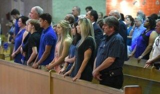 People standing in rows inside a courtroom, facing forward at a wooden railing.