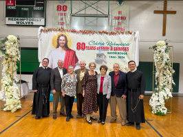 Group of people posing on a church gym floor before a banner reading “80 years of faith & service.”