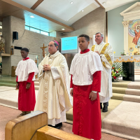 Clergy and altar servers in red and white robes stand in a church sanctuary.