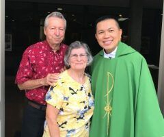 Three people posing together; one wears a green clergy robe, standing outdoors by a dark doorway.