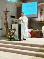 Priest in white vestments speaks at church altar beside crucifix and flowers.