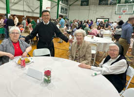 People seated at a round table in a gymnasium, with a man standing behind them smiling.