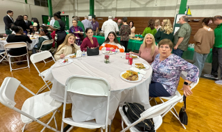 People seated at round tables in a gymnasium-style banquet hall, smiling during a gathering