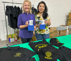 Two women hold blue mugs behind a table with black T-shirts and green tablecloth in a room.