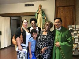 Five people posing indoors beside a statue of the Virgin Mary, one person in a green robe.