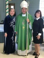 Three people posing indoors, with a man in green clerical robes between two women in black outfits.