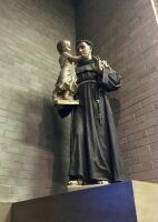 Statue of a robed figure holding a child, mounted on a pedestal against a brick wall.