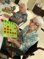 Two seniors playing bingo at a table, smiling and holding cards in a bright room.