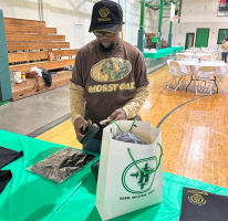 Person at a table opening a white gift bag in a gymnasium with green table covers and bleachers.