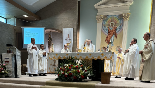 Priests standing at a church altar during Mass, with candles, flowers, and a religious mural behind them