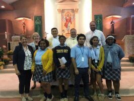 Group of students and adults posing in a church sanctuary, with a mural and wall sconces in the background