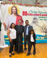 Four people posing indoors before a colorful church banner with a portrait and “80 years of faith & service” text