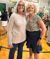 Two women smiling indoors, standing together at an event with artwork in the background.