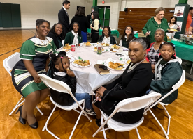 Group seated around a round table at a community gathering in a gymnasium, chatting over food.