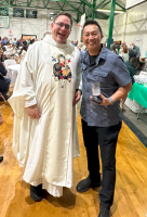 Two people smiling on a gym floor, one in a cream robe and the other in a gray shirt holding a camera.