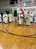 People in white robes stand on a gym floor during a religious ceremony with a banner in the background