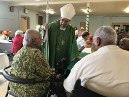 Clergyman in green vestments speaking with seated seniors in a community dining hall