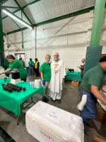 Indoor event booth with people serving green-covered tables and a robed man standing near the center