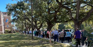People walking in a shaded park path beside a brick building, lined with trees and grass.