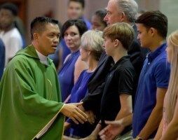 Priest in green vestments greets people standing in line indoors with handshake