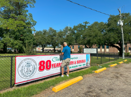 Person placing a banner on a chain-link fence beside a parking lot and trees