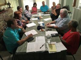People seated around a dining table in a living room, having a discussion with papers and notebooks.