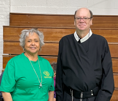 Two people standing in front of wooden bleachers, one in a green T-shirt and one in a black clerical shirt.