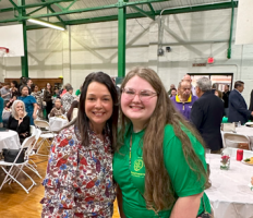 Two women smile indoors at a busy event, one in a floral blouse and one in a green shirt.