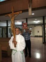 Man in white robe holding a crucifix in a hallway, with another man standing behind him.