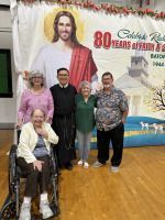Five people pose before a banner at a 80th anniversary celebration, one seated in a wheelchair.