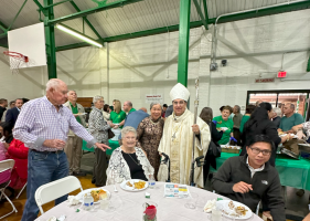 People gathered at a banquet table in a gymnasium, with one person in ceremonial white robes standing nearby.