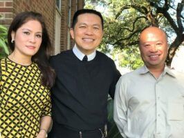 Three people posing outdoors, smiling in front of greenery and brick walls.