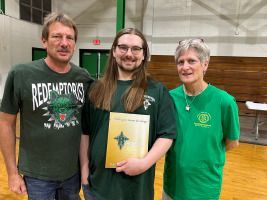 Three people stand indoors, smiling, with a certificate held by the person in the center.