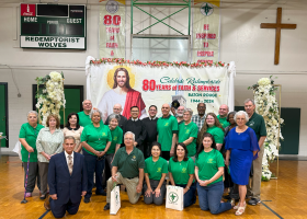 Group posing in a gym under an “80 years” banner, with a large religious banner and cross in the background.