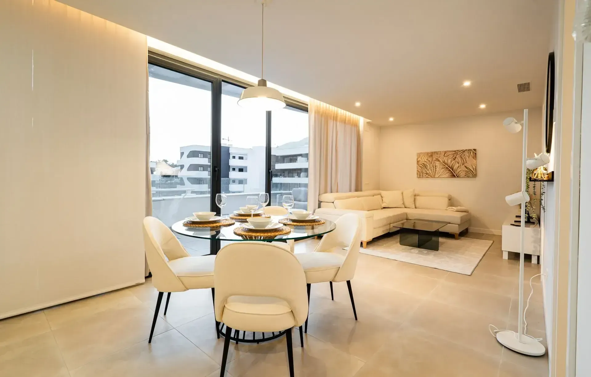Modern open-plan dining and living room with white furniture, sofa, and large windows overlooking city buildings