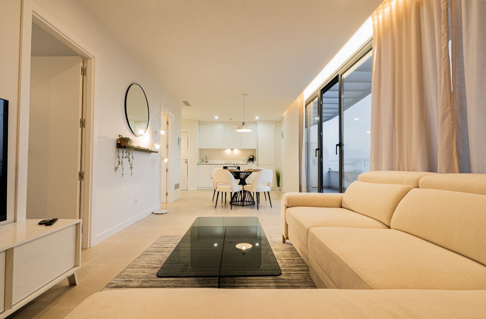 Modern white living room with beige sofa, glass coffee table, and open dining area by large windows