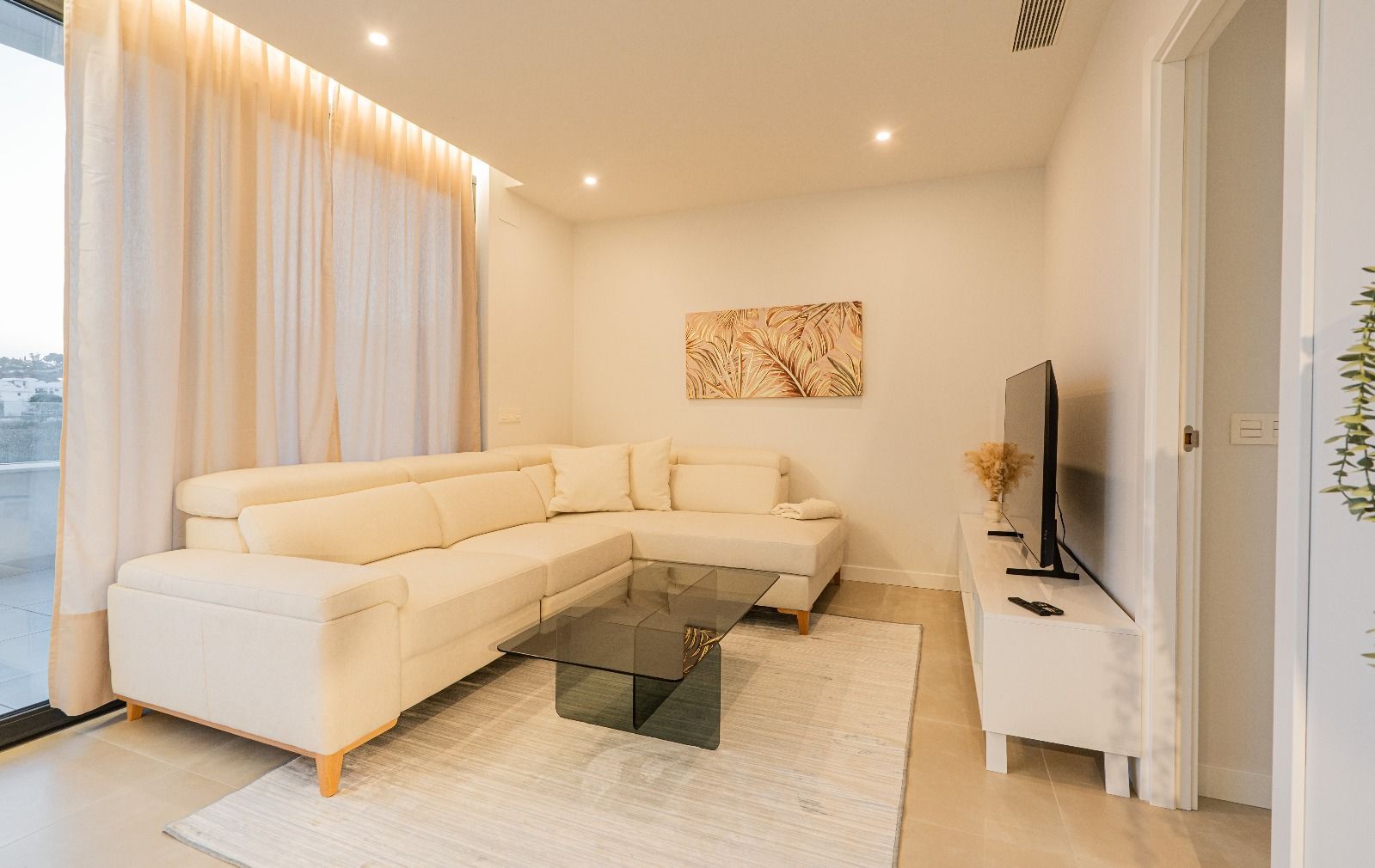 Bright modern living room with white sectional sofa, glass coffee table, TV, and floor-to-ceiling curtains