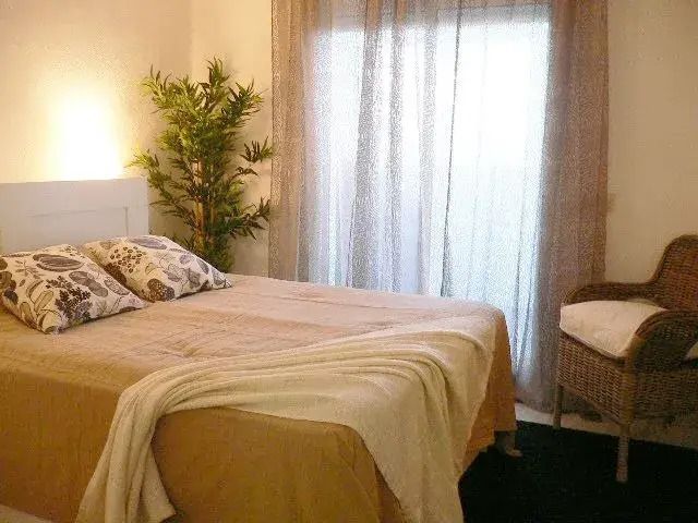 Cozy bedroom with a neatly made bed, patterned pillows, sheer curtains, and a wicker chair near a window